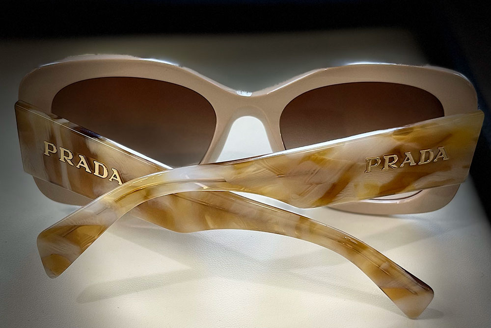 The image displays a pair of sunglasses with a tortoiseshell pattern, featuring the brand name 'Prada' prominently on the temples.