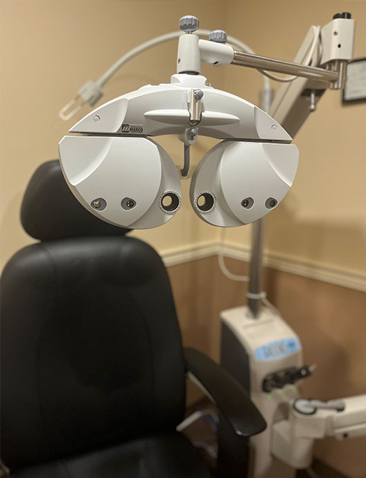 The image shows an optometry chair with a pair of binoculars attached to it, positioned in front of a desk and a chair, likely in an office setting.