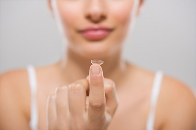 The image shows a close-up of a person s hand holding up a single contact lens against their finger, with the focus on the lens.