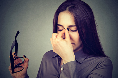 A woman in a grey blazer is holding her nose with one hand while looking down, with the other hand resting on her face  she appears to be in a state of distress or discomfort.