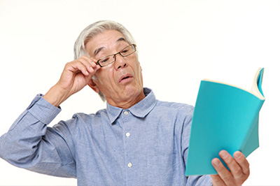 The image shows an older man with glasses, holding a blue book open and looking intently at the pages. He appears to be in a moment of discovery or concentration.