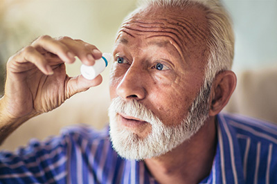 Elderly man with a white beard and mustache closely examining his eye with an ophthalmoscope.