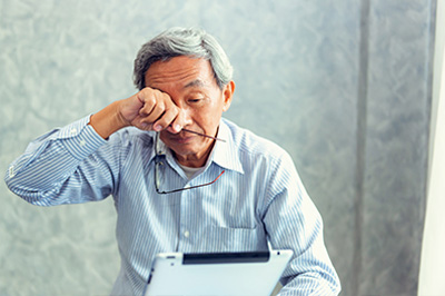 The image shows an older man with glasses, sitting at a desk and using a laptop. He appears to be in a professional setting, possibly an office or a study room.