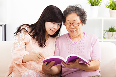 An elderly woman and a younger woman seated on a couch, both engrossed in reading a book together.