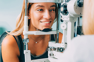 An image of a woman sitting in front of an eye exam machine, with a focus on her smiling face and the equipment behind her.