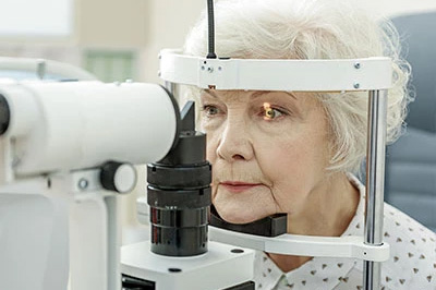 An elderly woman is seated at an eye examination machine, with a magnified view of her eyes through the device.