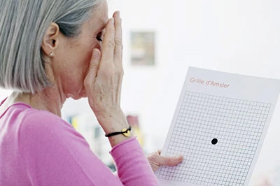 The image shows an older woman with her hand covering her face, possibly in a gesture of surprise or disbelief. She is looking at a piece of paper that appears to be a crossword puzzle or some kind of grid-based activity with text and numbers on it.